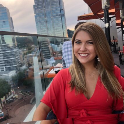 A woman in a red dress sits at an outdoor restaurant with city buildings in the background, smiling at the camera.