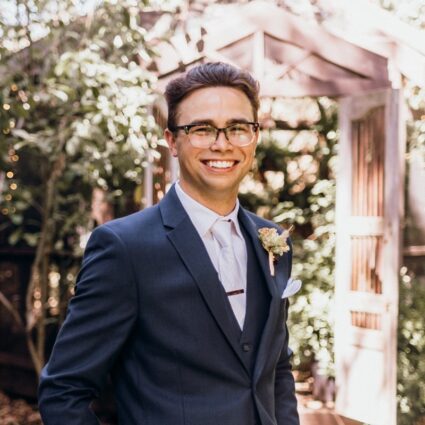 A man wearing a navy suit, white shirt, and glasses stands outdoors in front of a wooden structure, smiling. He has a boutonniere pinned to his lapel.