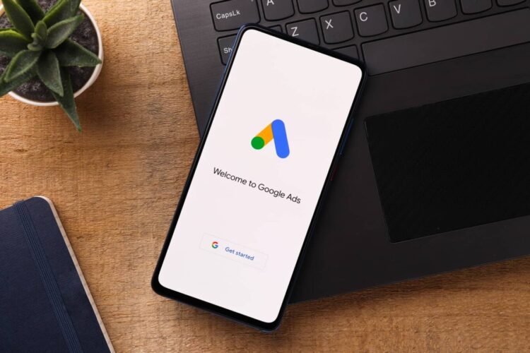 A smartphone on a wooden desk displays the Google Ads welcome screen, next to a laptop keyboard, a notebook, and a small potted plant.