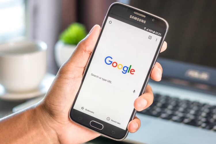 A person holds a Samsung smartphone displaying the Google search homepage, with a laptop and a cup in the blurred background.