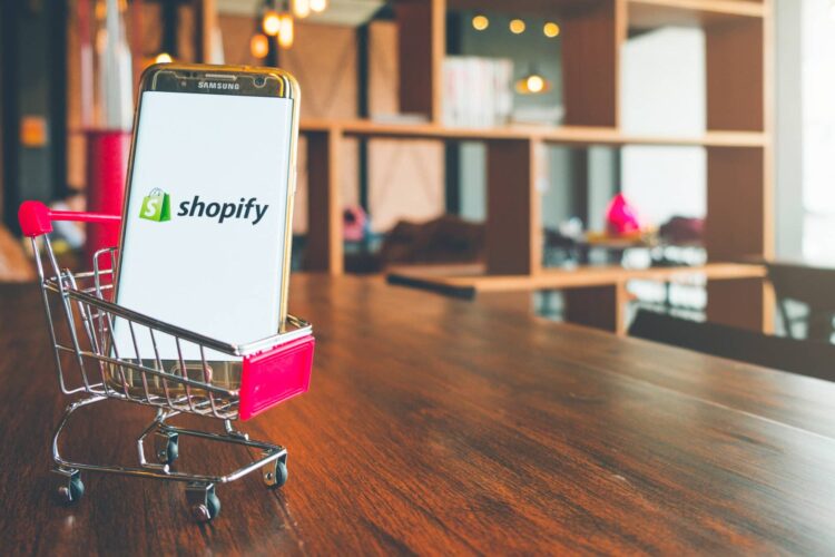 A smartphone displaying the Shopify logo sits inside a small shopping cart on a wooden table in a modern room.