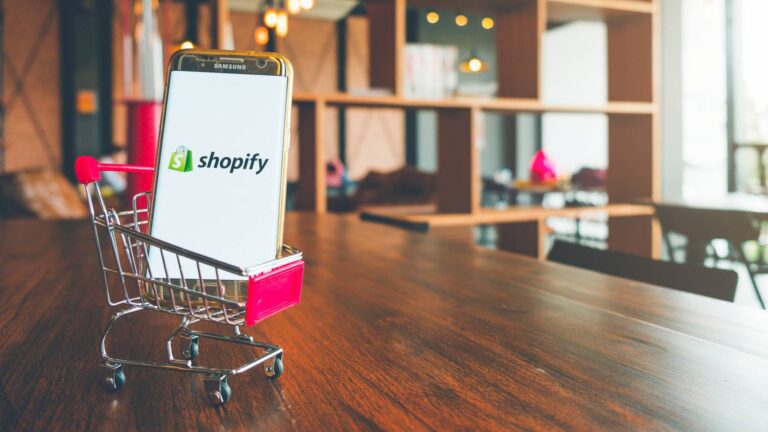 A smartphone displaying the Shopify logo sits inside a small shopping cart on a wooden table in a modern room.