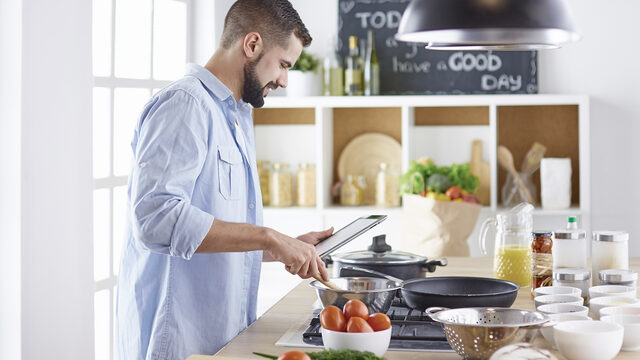 A man stands in a kitchen, cooking at the stove while looking at a tablet. Various ingredients and cooking utensils are arranged on the counter.