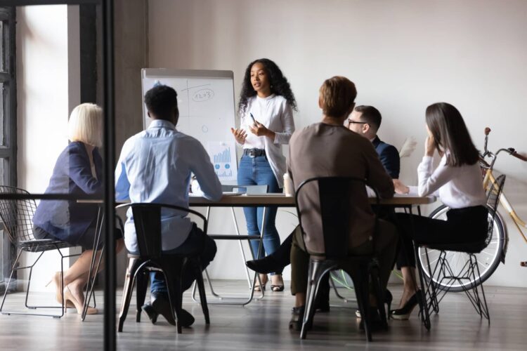 A woman stands and presents to five seated colleagues in a modern office meeting room with a flipchart and a bicycle in the background.