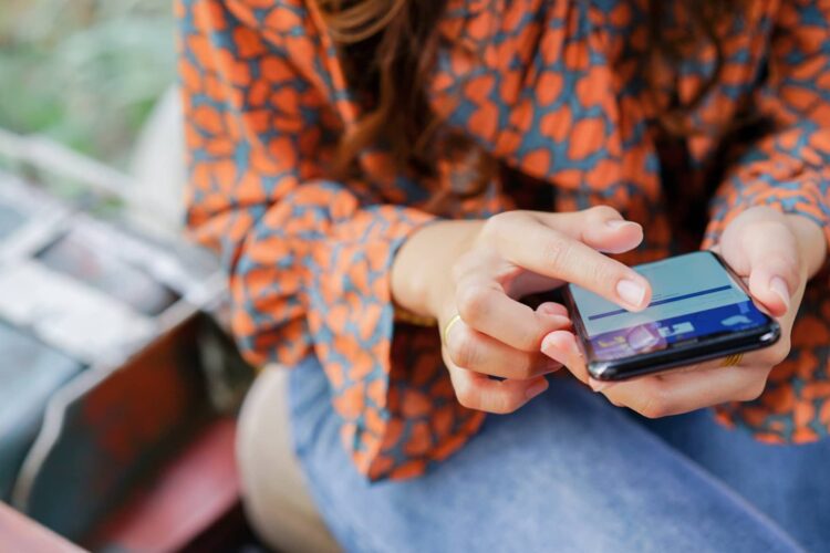 A person wearing a patterned orange blouse uses a smartphone, scrolling with their index finger while sitting outdoors.