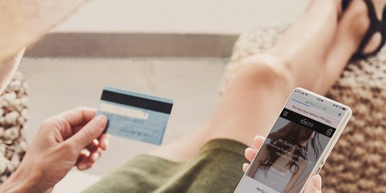 A person sitting outdoors holds a credit card and uses a smartphone for online shopping while wearing a wide-brimmed hat.
