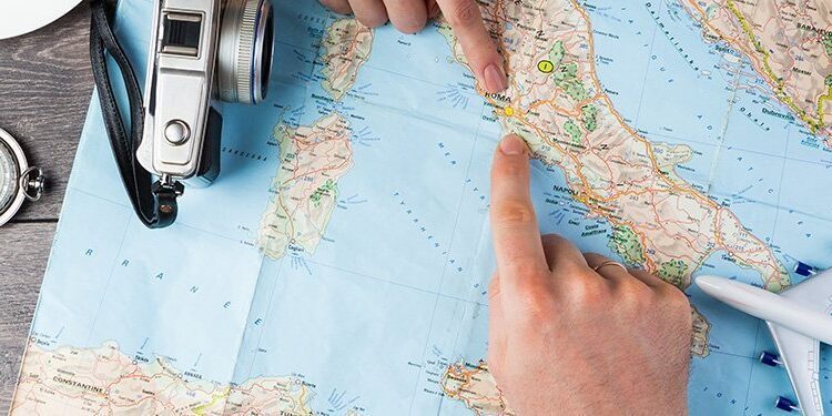 Two hands point at locations on a map surrounded by a camera, compass, toy airplane, and coffee cup on a wooden surface.