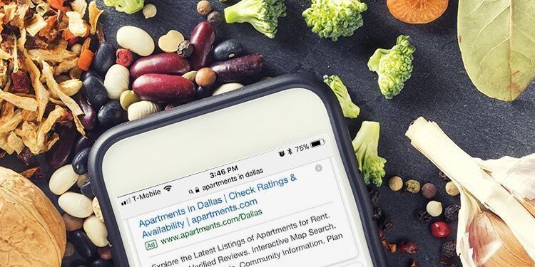 A smartphone displaying a health article is placed among assorted dried herbs, beans, garlic, bay leaves, and a wooden spoon on a dark surface.