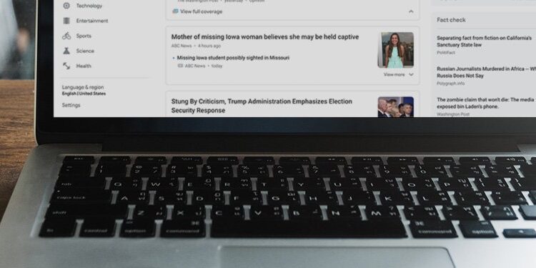 A laptop displaying a news website sits on a wooden table next to a glass cup of coffee and a saucer with a spoon.