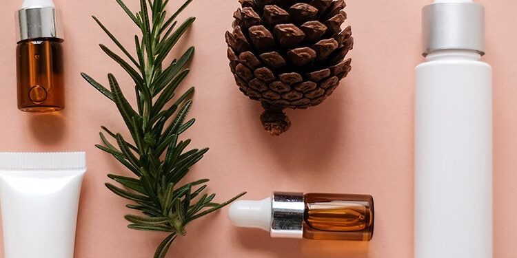 Skincare product bottles, a pine cone, a sprig of rosemary, and green leaves arranged on a light pink background.