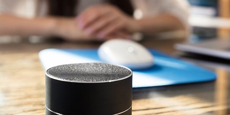 A smart speaker sits on a desk in the foreground, with a person, computer mouse, and laptop blurred in the background.