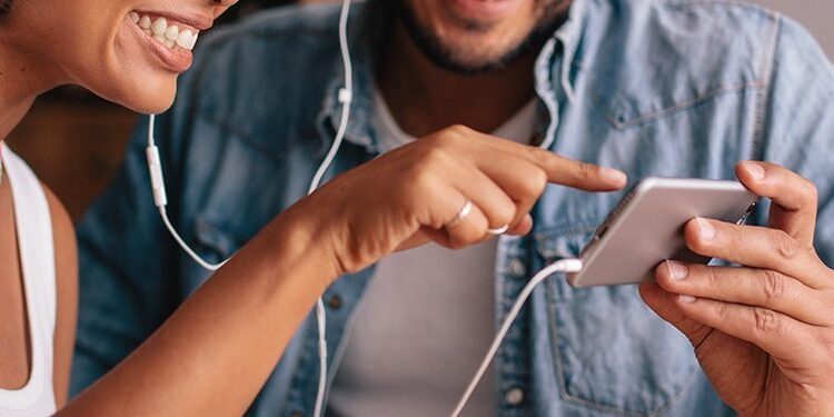 Two people wearing earbuds share a smartphone, smiling and looking at the screen together.
