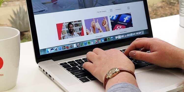 Person typing on a laptop at a desk with a mug, potted plant, and glass of water; social media images displayed on the laptop screen.