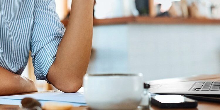 Person sitting at a wooden table with a laptop, a cup of coffee, papers, and a phone, in a well-lit indoor setting.