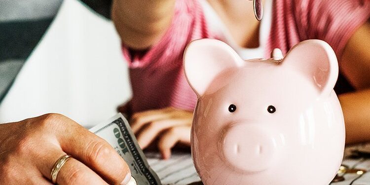 Three people sit at a table, placing money into a pink piggy bank, with loose cash and coins scattered nearby.