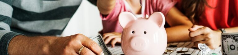Three people sit at a table, placing money into a pink piggy bank, with loose cash and coins scattered nearby.