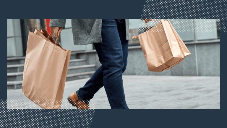 Person walking on a city sidewalk carrying brown paper shopping bags in both hands, wearing a gray coat, blue jeans, and tan shoes.