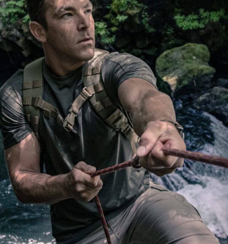 A man wearing a backpack and t-shirt climbs with a rope beside a flowing stream in a forested area.