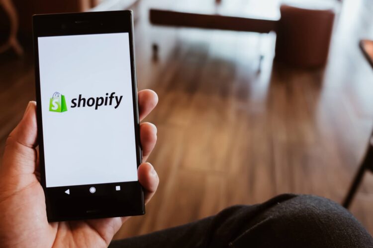 A person holds a smartphone displaying the Shopify logo on the screen, with a blurred indoor background.
