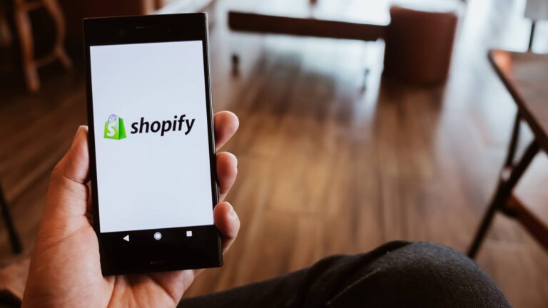 A person holds a smartphone displaying the Shopify logo on the screen, with a blurred indoor background.