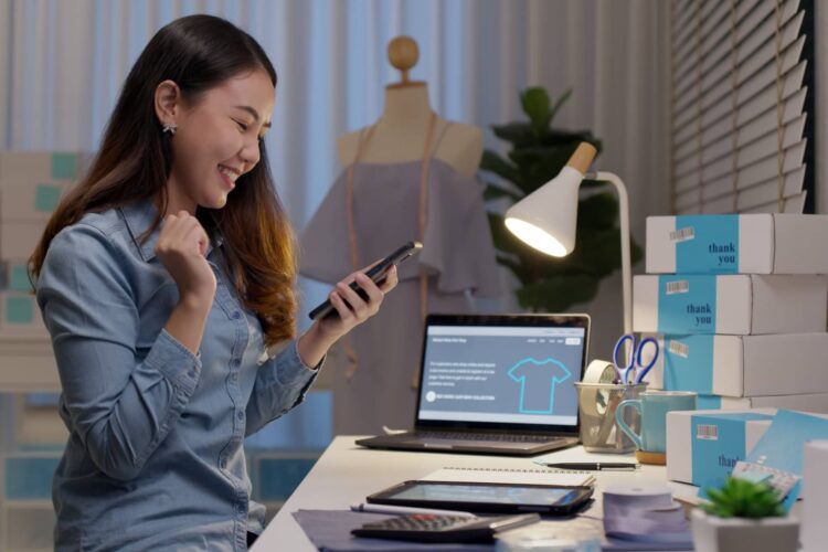 A woman sits at a desk with packaging boxes, a laptop, and a smartphone, appearing pleased in a workspace, possibly managing an online shop.