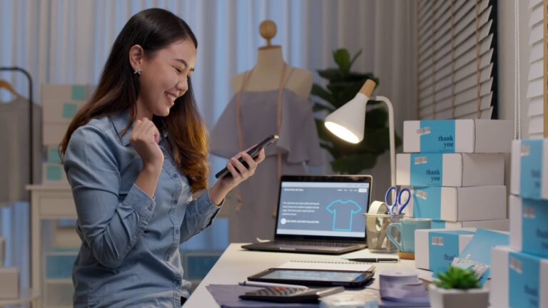 A woman sits at a desk with packaging boxes, a laptop, and a smartphone, appearing pleased in a workspace, possibly managing an online shop.