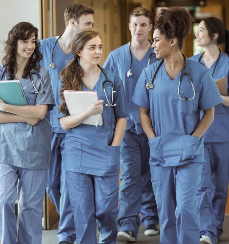 A group of young medical professionals in blue scrubs walk together in a hallway, holding notebooks and wearing stethoscopes.