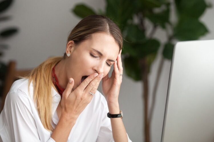 A woman sitting at a desk in front of a computer yawns and rests her head on one hand, appearing tired or fatigued.