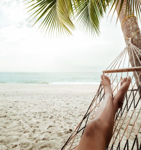Person lying in a hammock under a palm tree on a sandy beach, facing the ocean with calm waves and a cloudy sky in the background.