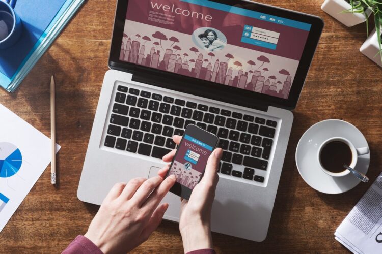 Person using a smartphone in front of an open laptop displaying a welcome screen, with a coffee cup, newspaper, and office supplies on a wooden desk.