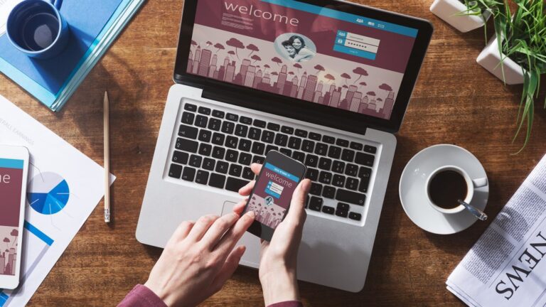 Person using a smartphone in front of an open laptop displaying a welcome screen, with a coffee cup, newspaper, and office supplies on a wooden desk.
