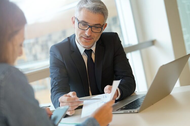 A man in a suit reviews documents with a woman at a desk, with a laptop and papers in front of them in a modern office setting.