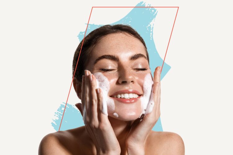 A woman with a bare shoulder smiles with closed eyes while applying foamy facial cleanser to her cheeks, against a white background with blue and pink graphic accents.