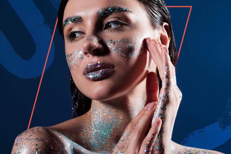 A woman with wet hair poses with her hands near her face, skin and lips covered in silver and blue glitter, against a blue geometric background.