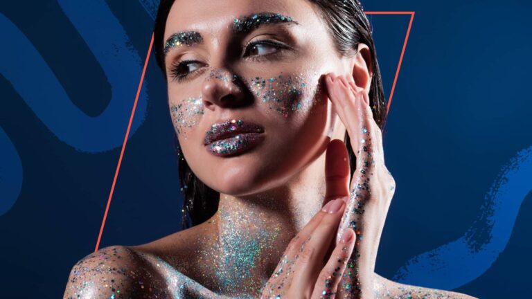 A woman with wet hair poses with her hands near her face, skin and lips covered in silver and blue glitter, against a blue geometric background.