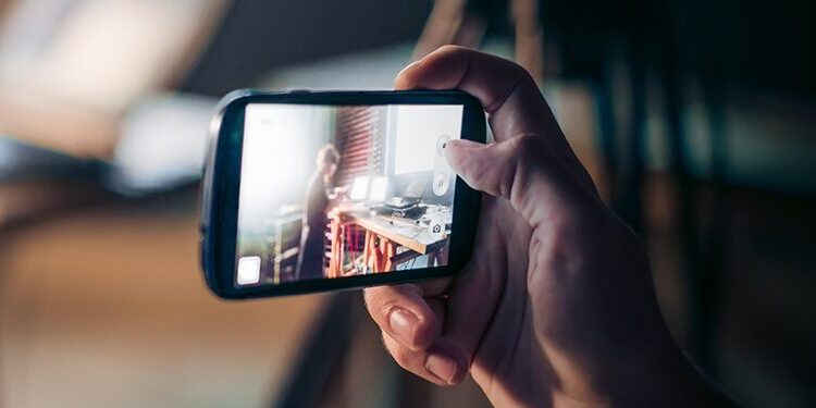 Close-up of a hand holding a smartphone, capturing a photo or video of a person sitting at a desk with electronic equipment in a softly lit room.
