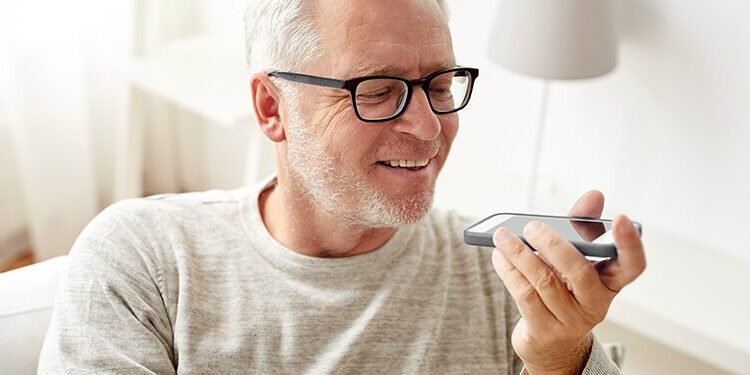 Older man with glasses sits indoors, holding a smartphone and speaking into it, appearing to use a voice assistant feature.