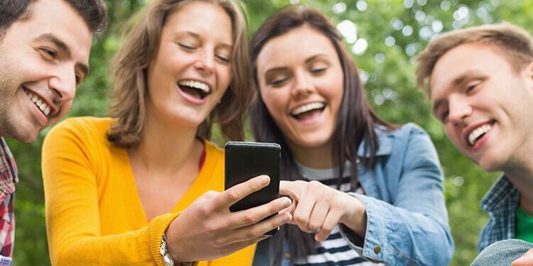 Four young adults sitting outdoors and looking at a smartphone together, smiling and laughing, with trees in the background.