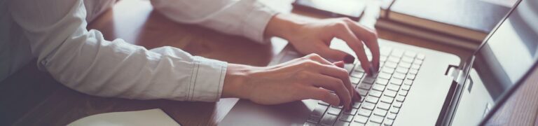 Person in a white shirt typing on a laptop keyboard at a wooden desk with notebooks nearby.