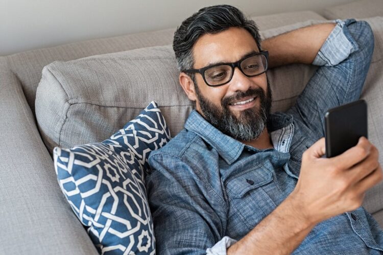 Man with glasses and a beard reclines on a couch, smiling while looking at his smartphone. He is wearing a denim shirt and has a patterned cushion behind him.