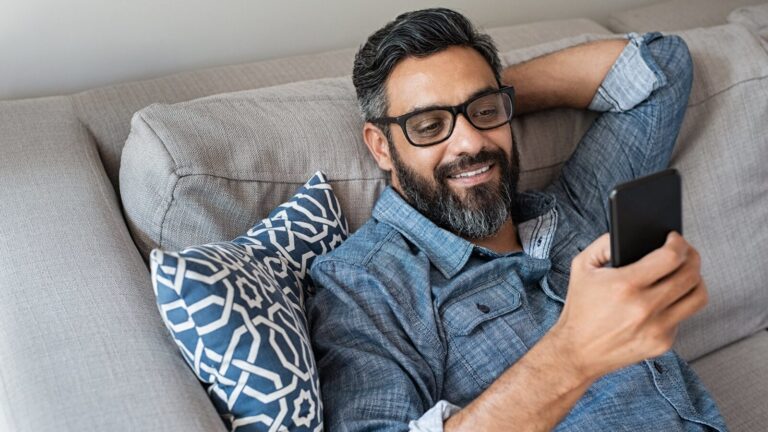 Man with glasses and a beard reclines on a couch, smiling while looking at his smartphone. He is wearing a denim shirt and has a patterned cushion behind him.