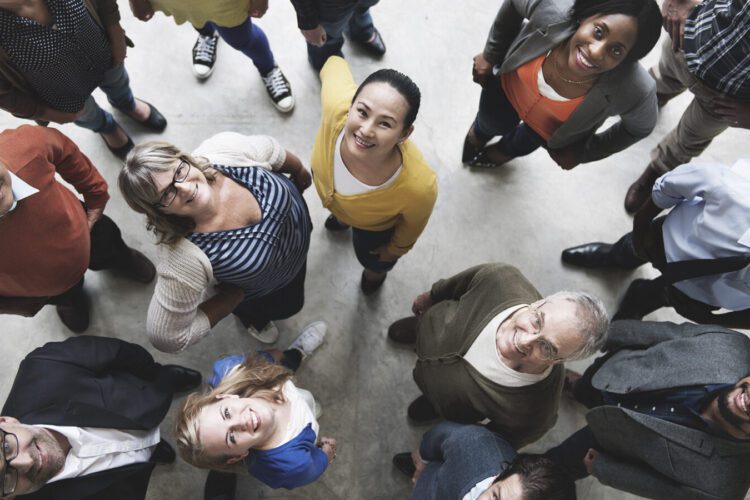 A diverse group of people standing in a circle, looking up toward the camera, photographed from above on a light-colored floor.