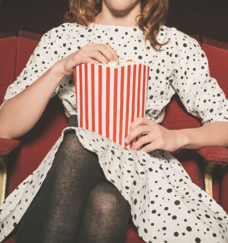 A person in a polka dot dress sits in a red theater seat, holding a large striped popcorn container and eating popcorn.