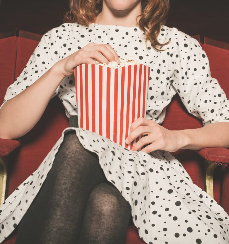 A person in a polka dot dress sits in a red theater seat, holding a large striped popcorn container and eating popcorn.