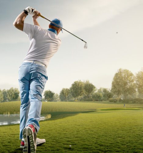 A golfer in mid-swing watches the ball fly down a fairway on a sunny golf course, with trees and a pond in the background.
