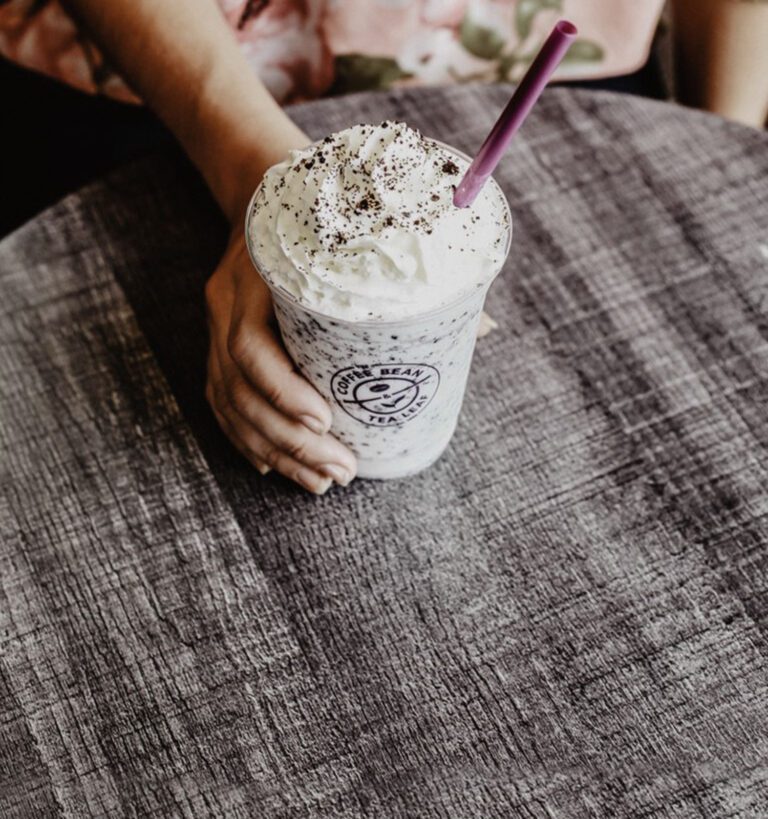 A person holds a whipped cream-topped iced blended drink with chocolate sprinkles and a purple straw on a gray textured table.