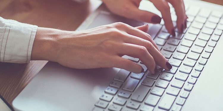 A person wearing a white shirt types on a silver laptop keyboard, with an open notebook and pen nearby on a wooden desk.