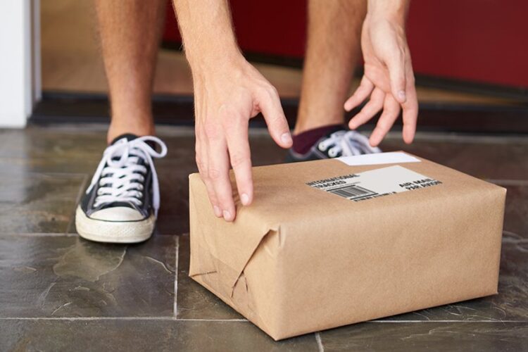 Person wearing sneakers picking up a brown paper-wrapped package with a shipping label from a doorstep.