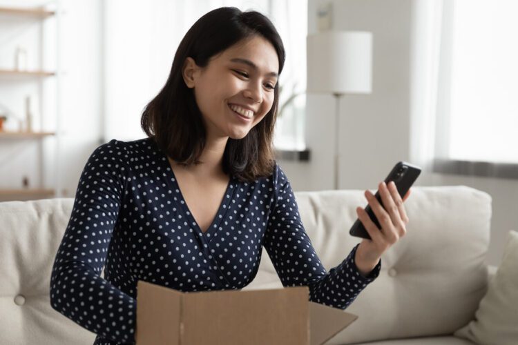 A woman sits on a couch, smiling at her smartphone while opening a cardboard box in a well-lit living room.