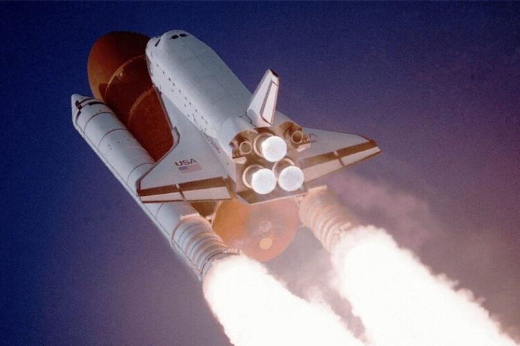 A space shuttle launches into the sky with rockets firing and flames visible against a clear blue background.